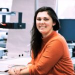 Photograph with a woman in an orange jumper and brown hair sat at a desk.